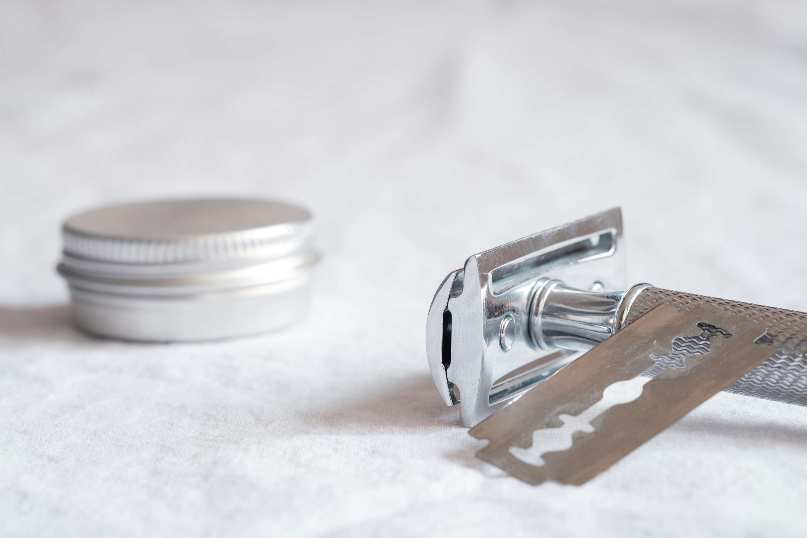 A metal safety razor and a loose blade resting on white linen beside a small aluminium tin