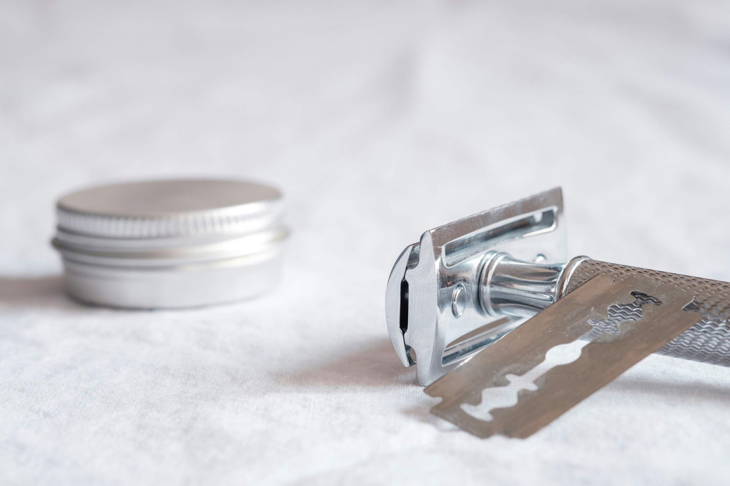 A metal safety razor and a loose blade resting on white linen beside a small aluminium tin