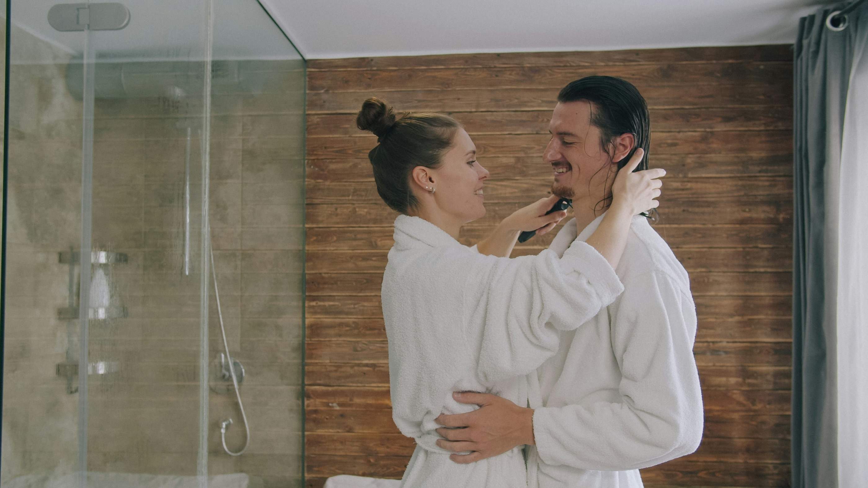 A couple in white bathrobes in a wood-panelled bathroom, one smoothing back the other’s damp hair