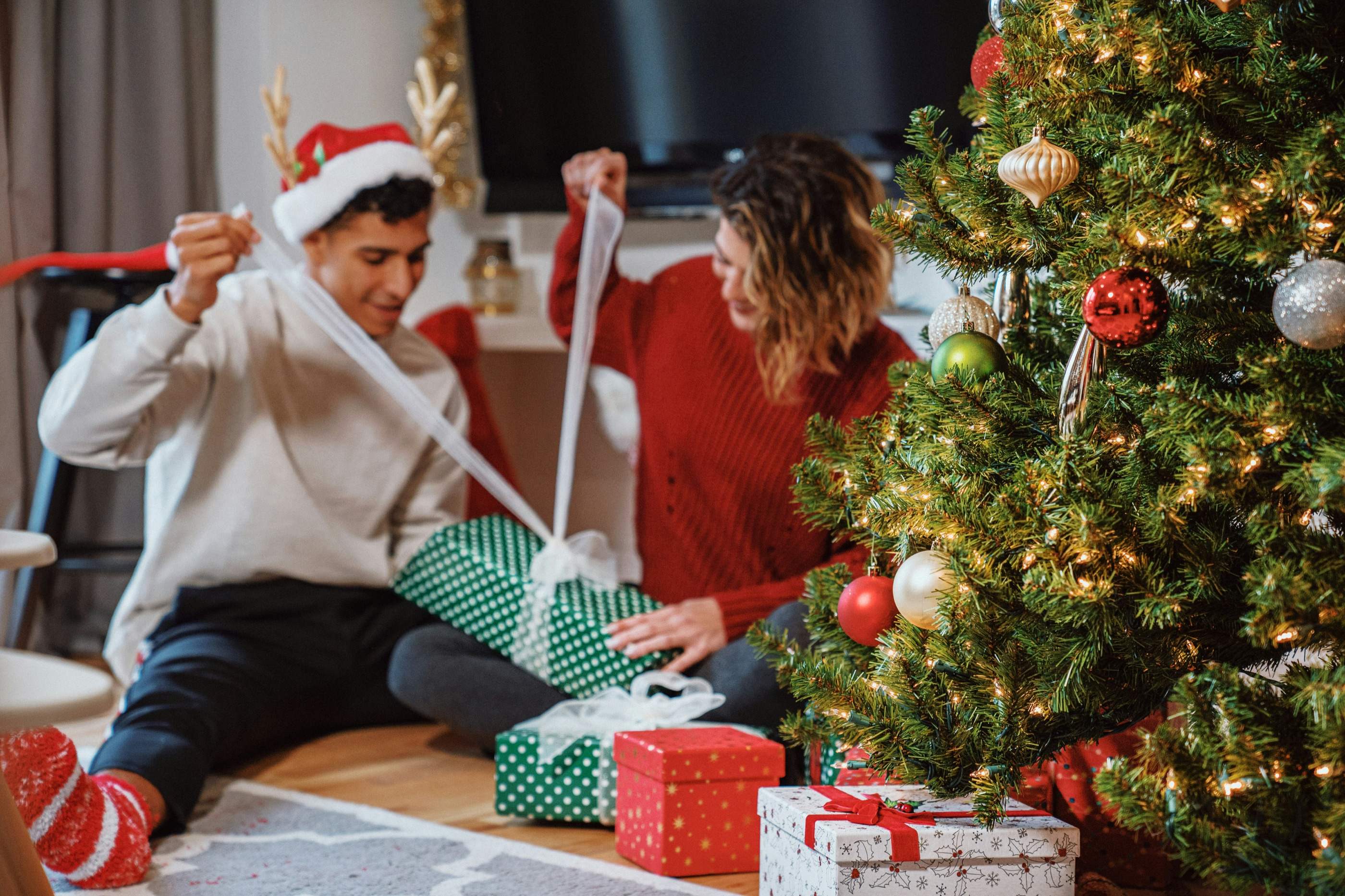 Two people unwrapping presents together on the floor beside a decorated Christmas tree