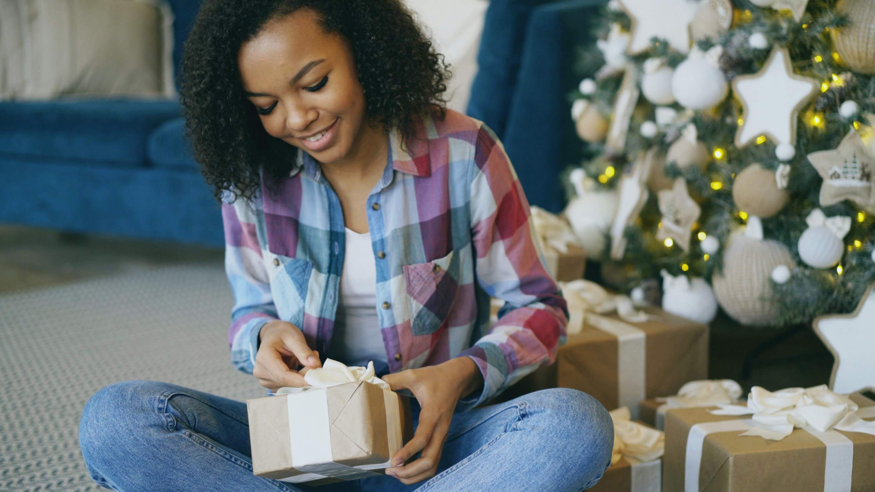 A woman sitting on the floor beside a Christmas tree, untying a kraft-paper gift