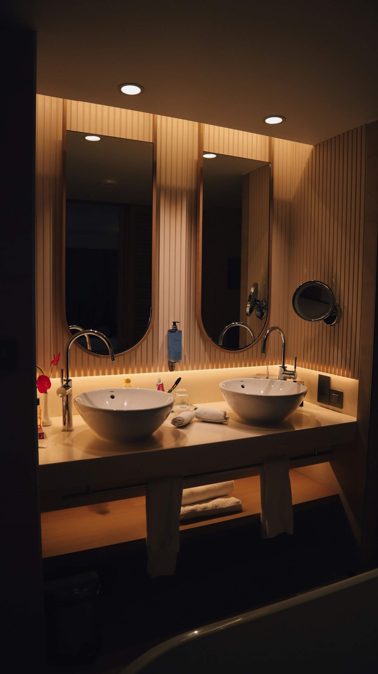 Two white vessel sinks under oval mirrors on a softly lit, wood-slatted bathroom vanity