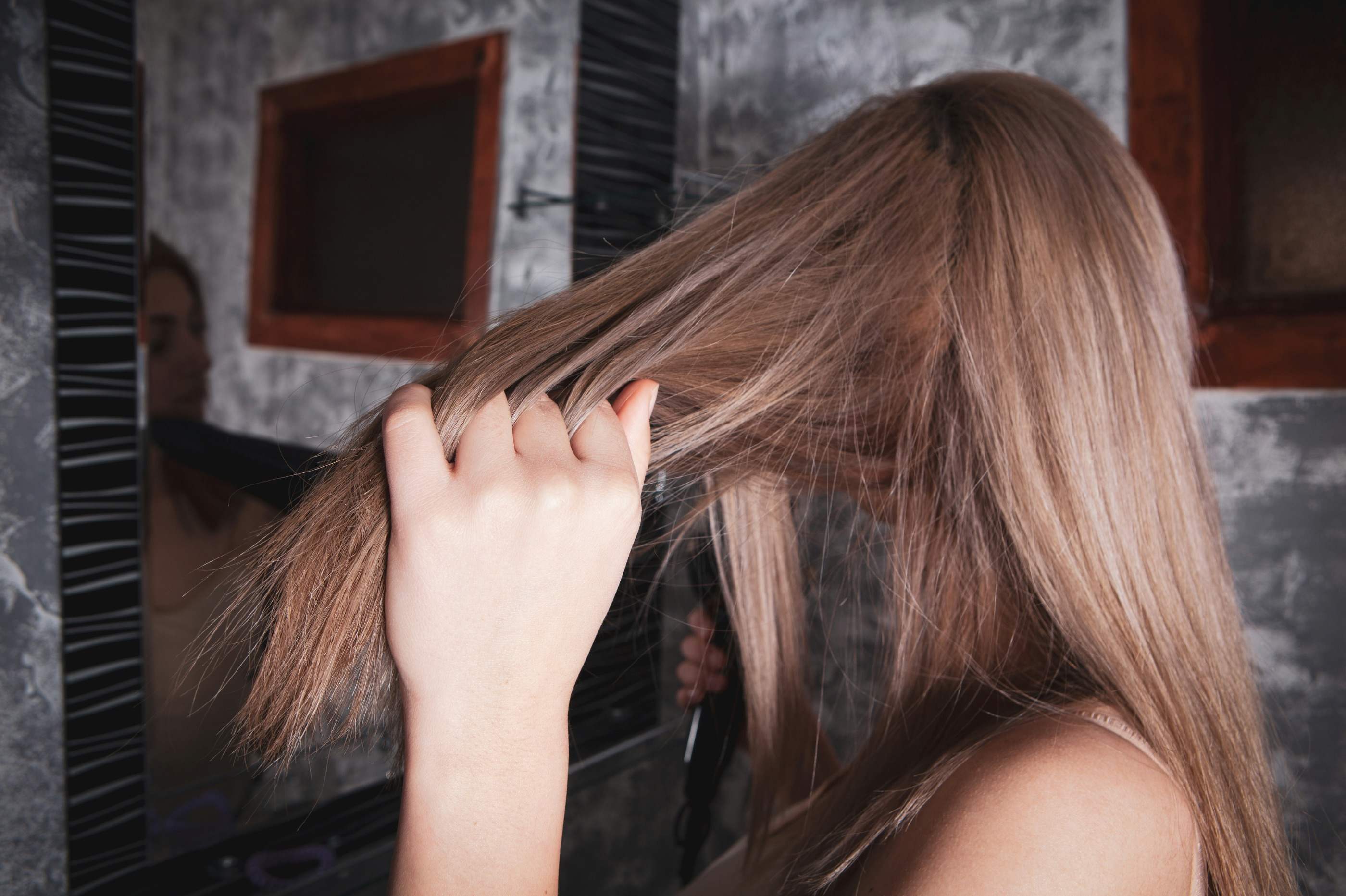 A woman running her fingers through long, straight hair in front of a bathroom mirror