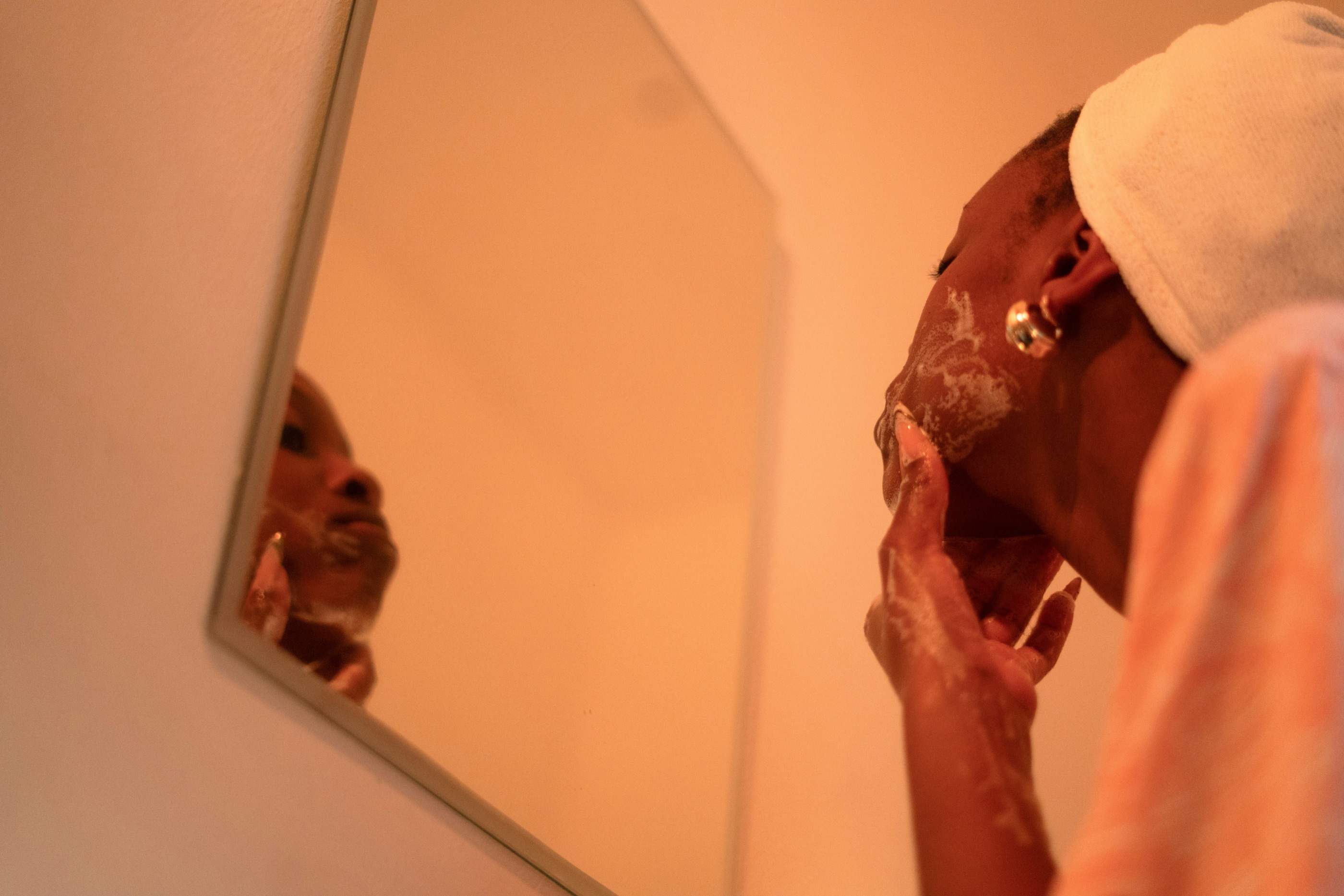 A man spreading shaving foam along his jaw while looking into a mirror, in warm orange light