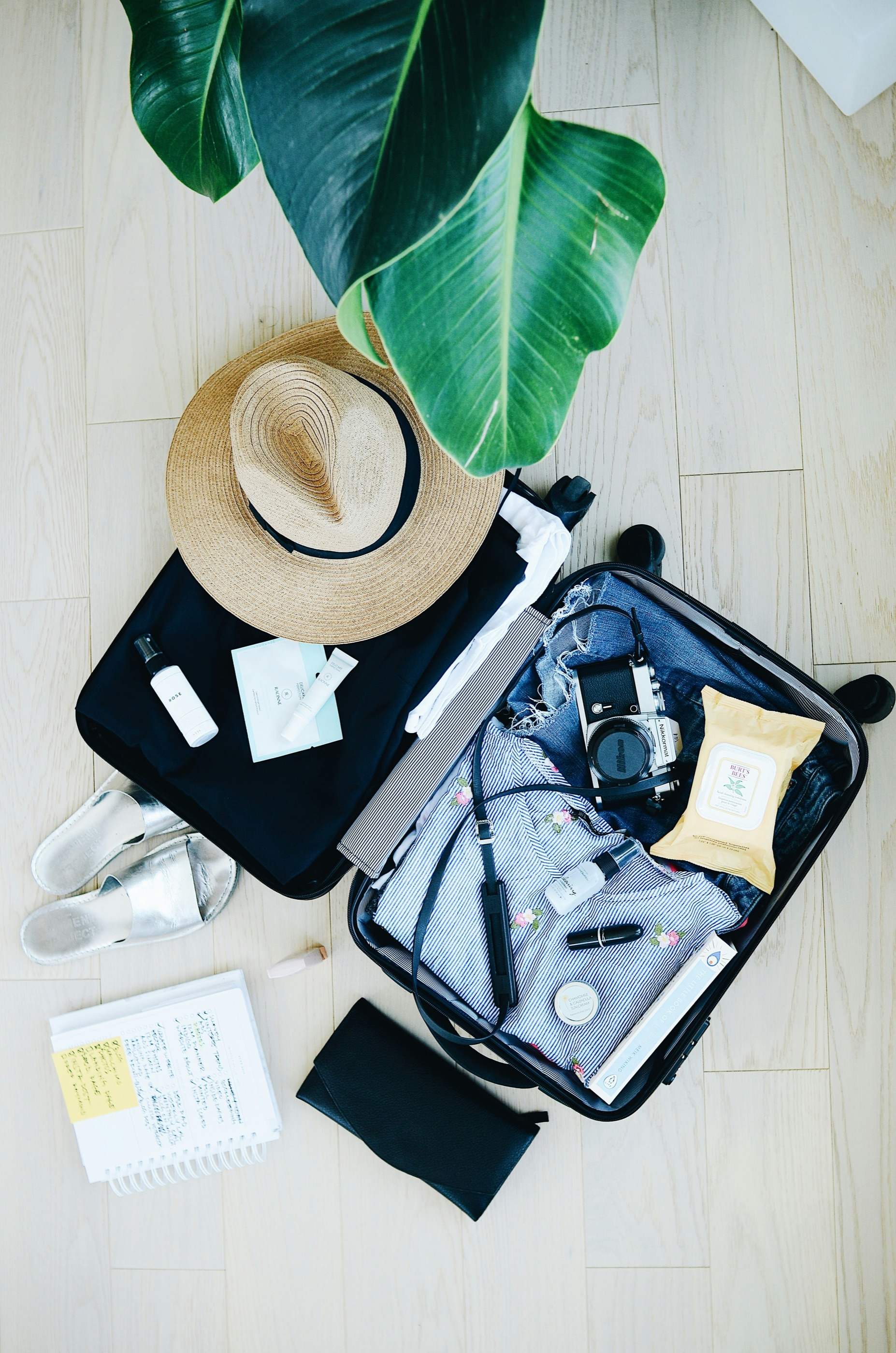 An open suitcase seen from above, packed with folded clothes, a camera and small pouches, with a straw hat resting on top